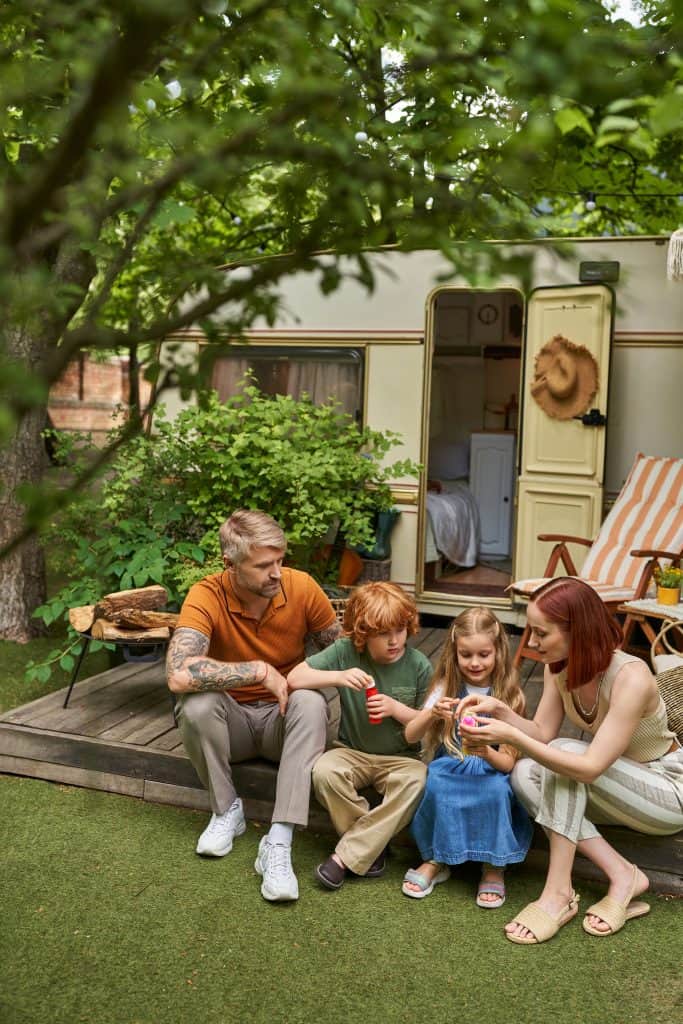 A family of four sits together on grass outside a camper van, sharing a moment and holding toys, with trees and outdoor furniture around them.