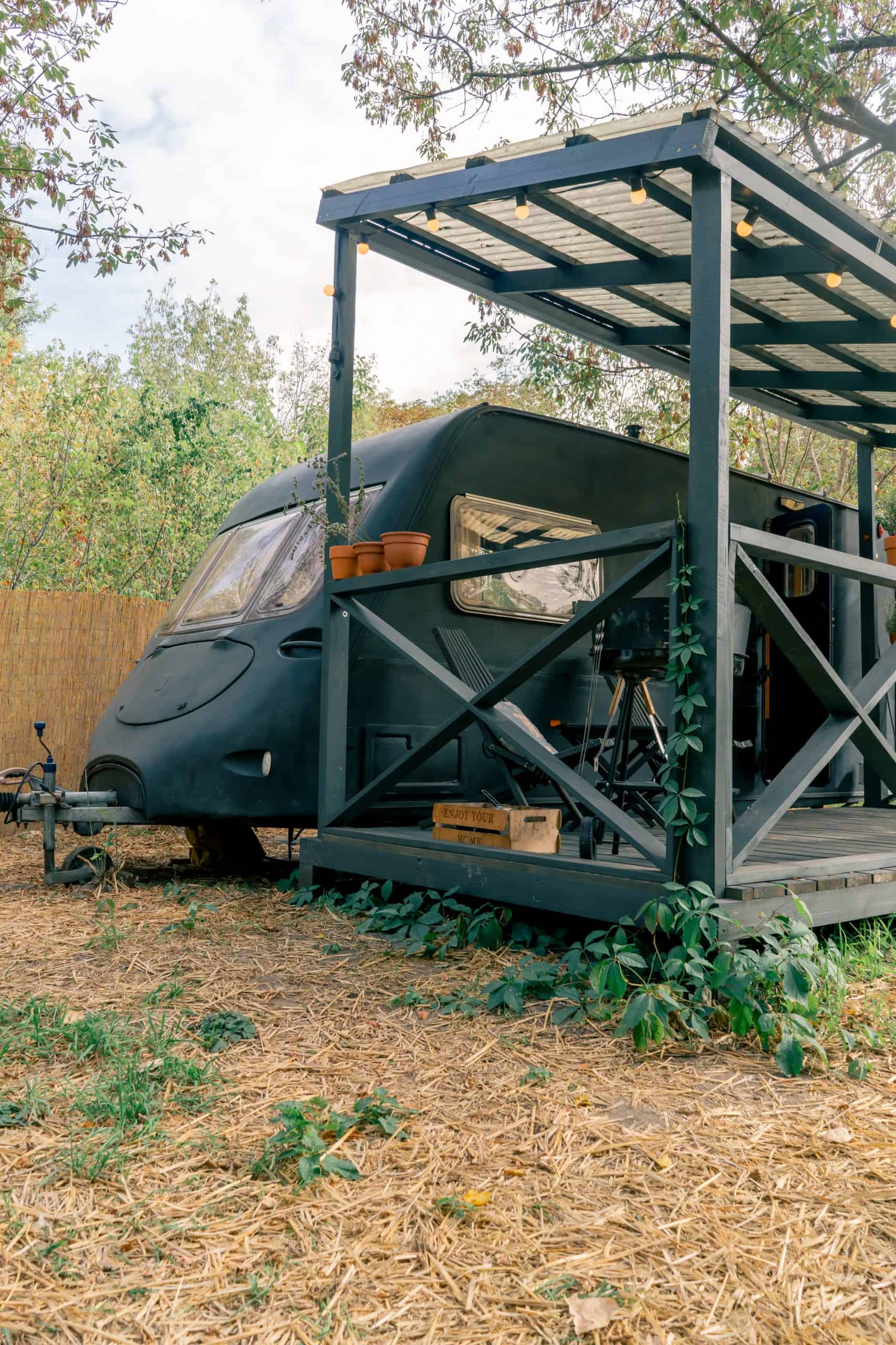 A black camper is parked next to a small wooden deck with potted plants, surrounded by trees and straw-covered ground.