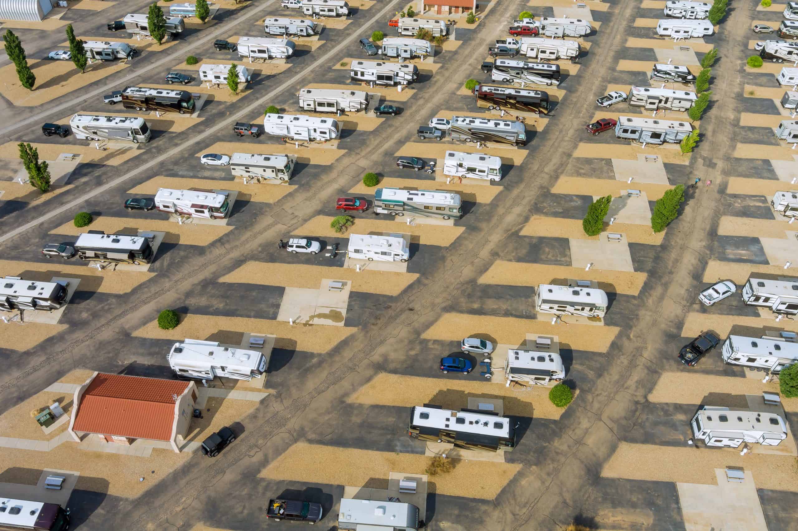 Aerial view of an RV park with rows of parked recreational vehicles, cars, and small buildings on gravel and paved lots, separated by trees.