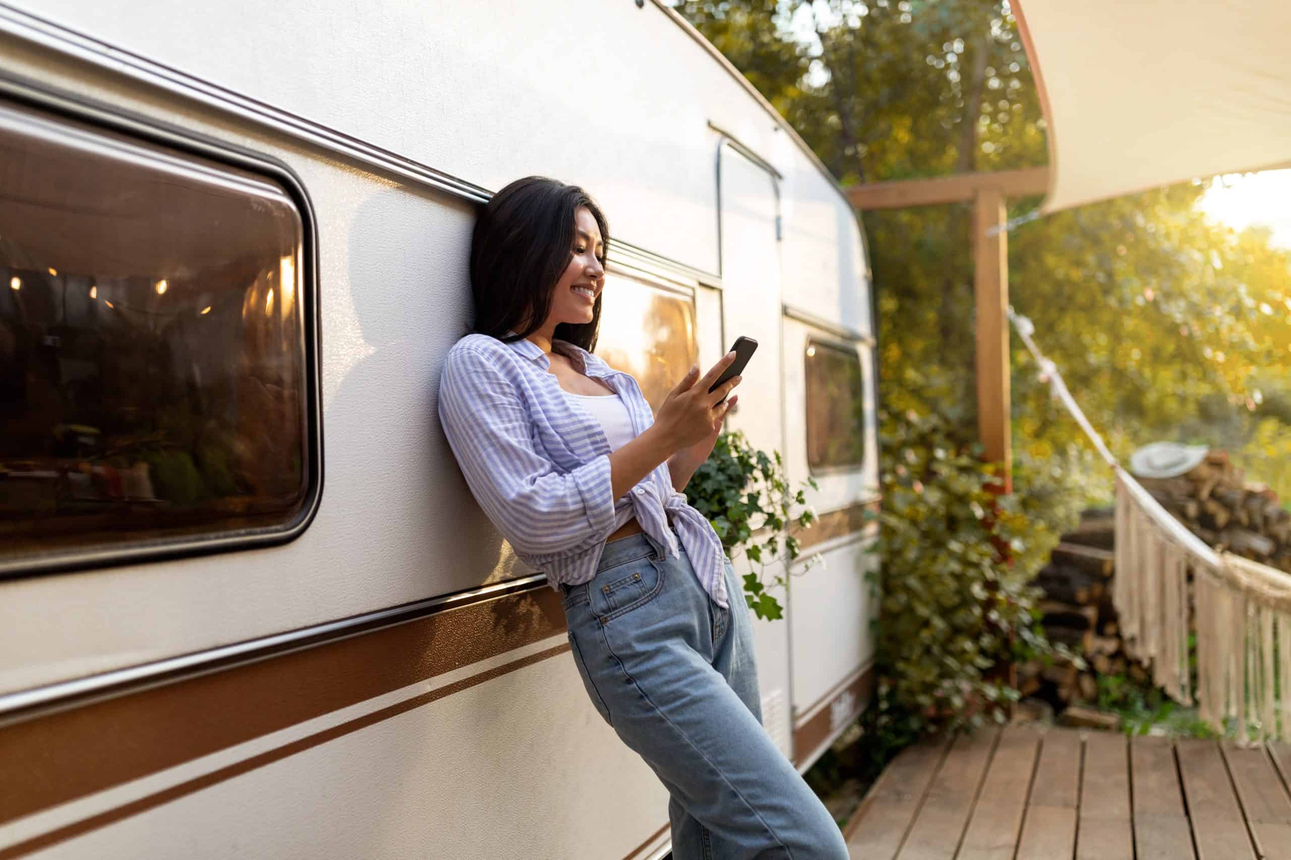 A woman leans against a camper, smiling while looking at her phone, with greenery and sunlight in the background.